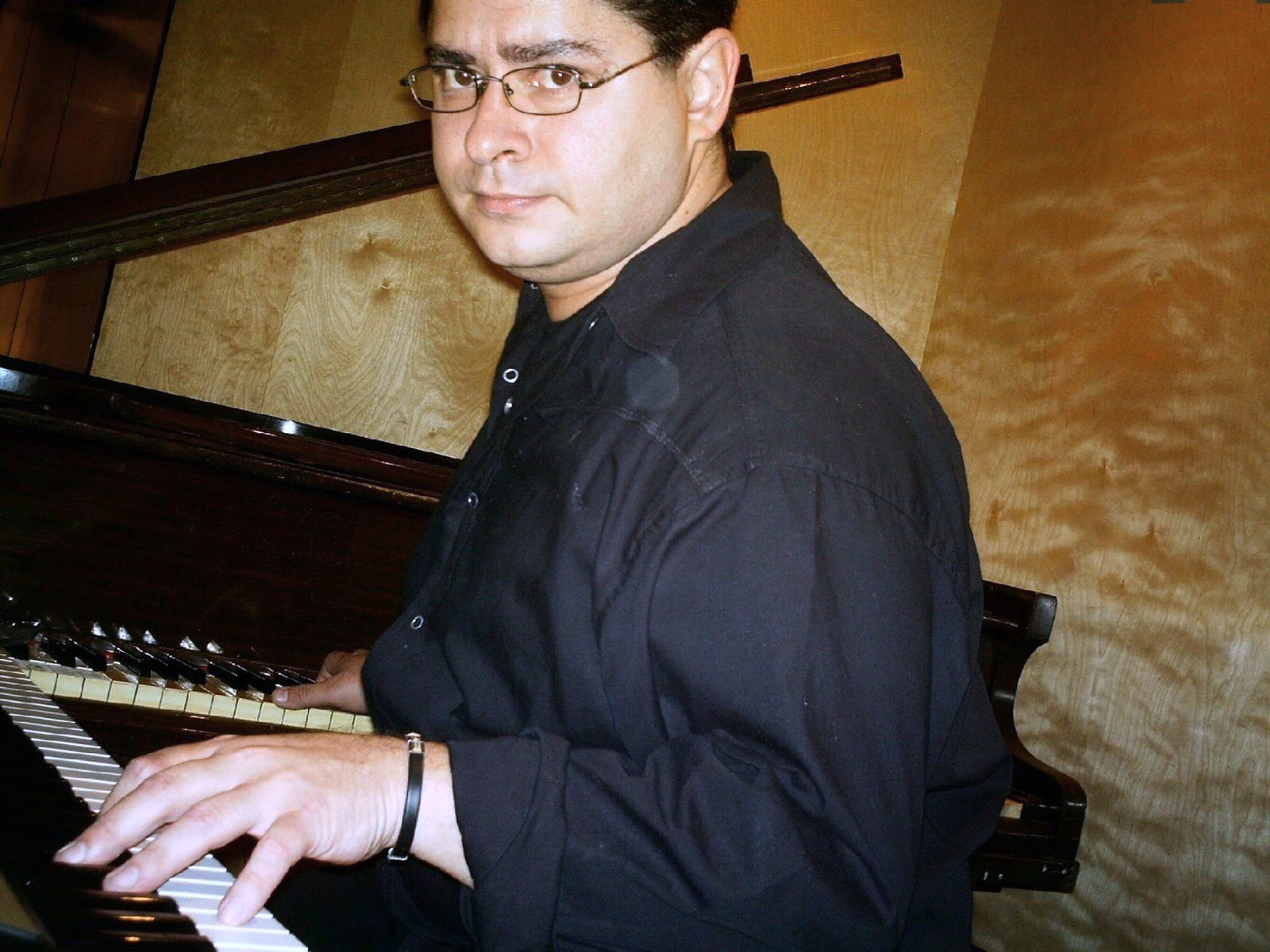 Yan Kayary con camisa negra y lentes, con las manos sobre un piano acústico