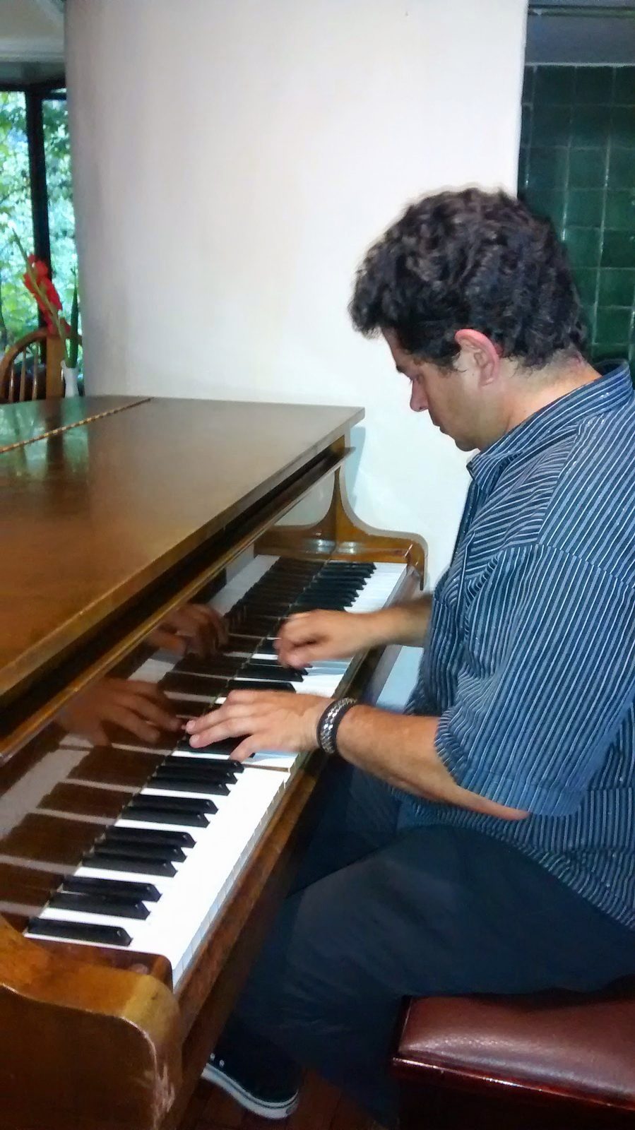 Yan Kayary con camisa azul, tocando un piano vertical de madera junto a una ventana