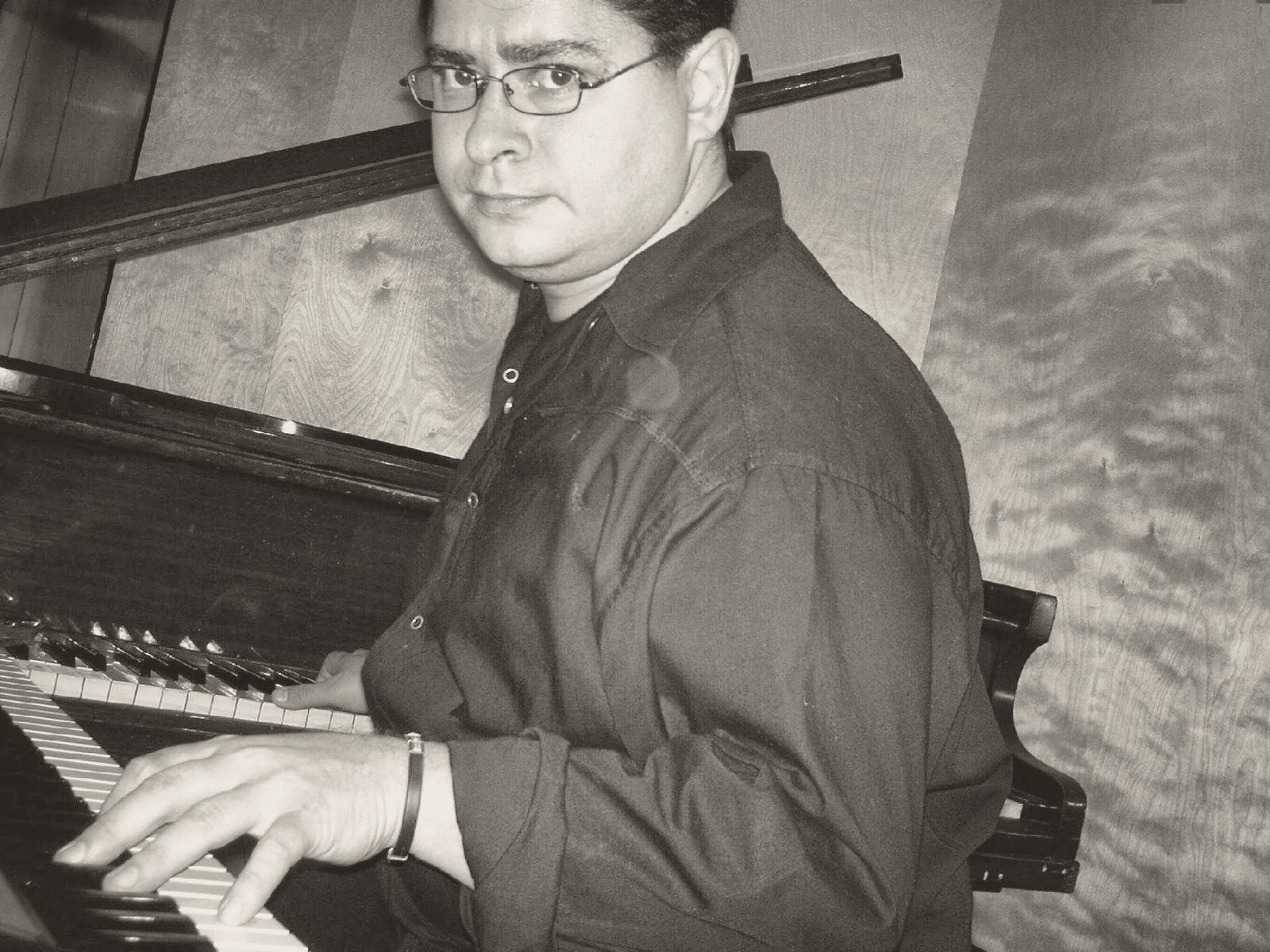 Yan Kayary con camisa negra y lentes, con las manos sobre un piano acústico